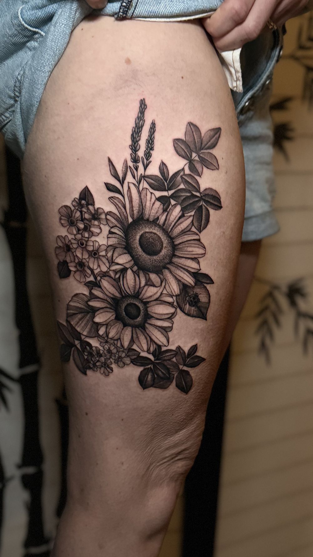 Black and white floral tattoo on a person's thigh, featuring sunflowers, lavender, and other flowers.
