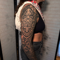 Woman with ornate black and gray sleeve tattoo, wearing a black top and jeans.