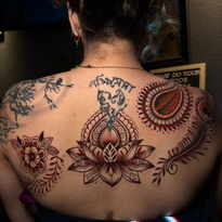 Back of person with multiple red and brown henna-style tattoos: flowers, feathers, a lotus, and circular designs.