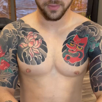 Man with upper body tattoos: lotus flower, red demon mask, and wave pattern in black, red, and pink.