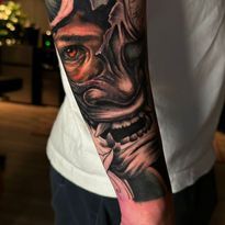 Tattoo of a demonic Japanese mask and eye on a forearm. Black, gray, and red ink.
