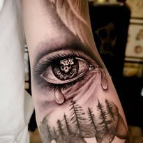 Realistic eye tattoo with tear rolling down, trees in background and skull in the iris.