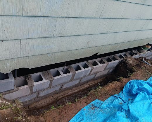 Damaged house siding above cinder blocks, with exposed soil and a blue tarp beside the foundation