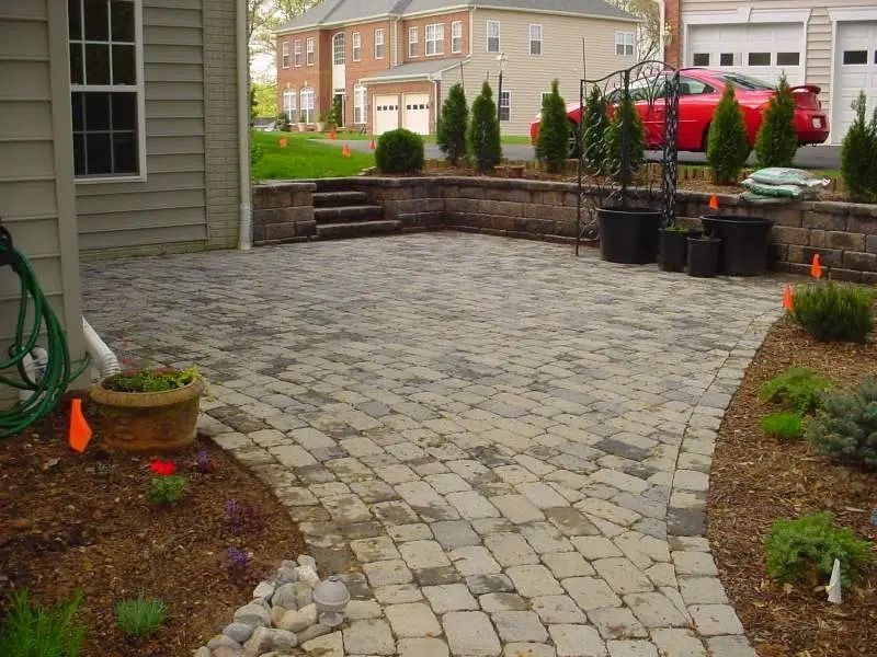 Stone paver patio beside a house, with potted plants, mulch beds, and a brick retaining wall in the background