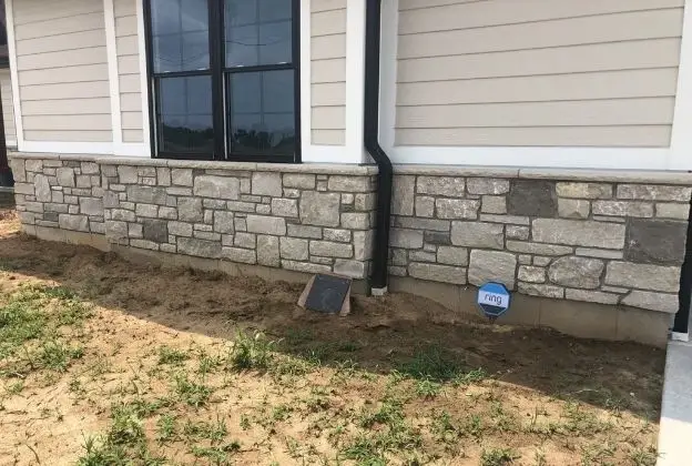 House exterior with stone foundation, beige siding, downspout, and a patchy dirt yard.