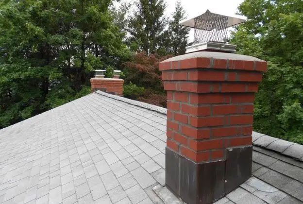 Brick chimney on a gray shingled roof, with trees in the background.