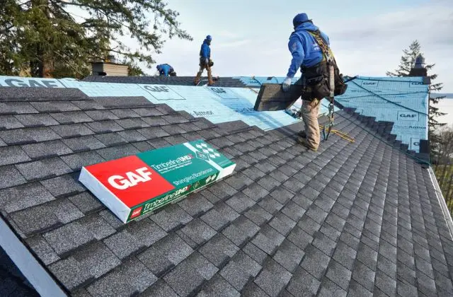 Roofers installing blue underlayment on a shingled roof, with a GAF roofing materials box in the foreground.