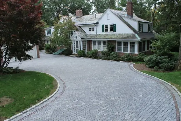 Curved brick driveway leading to a white house with a porch and trees surrounding it