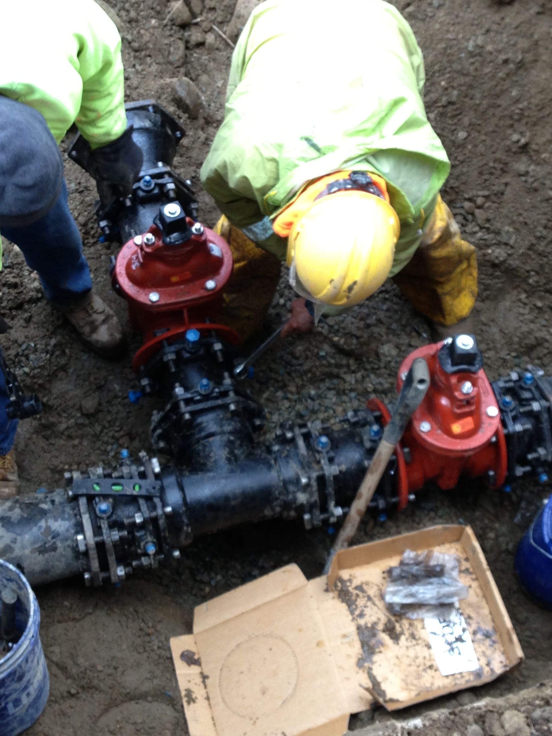 Workers in high-visibility gear repairing a buried water pipe with red valves in a muddy trench