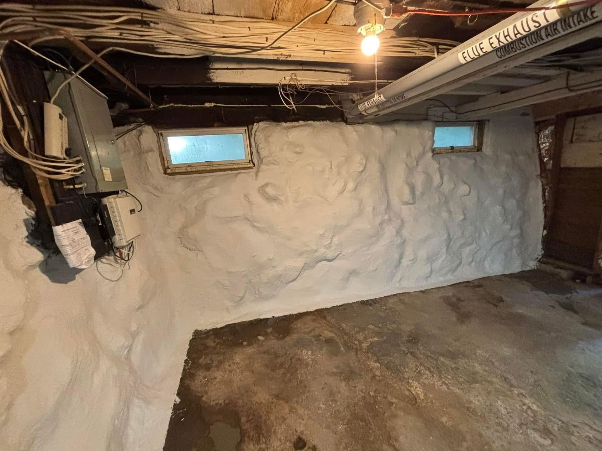 Basement with spray-foam insulated walls, small windows, exposed pipes, and a bare concrete floor.