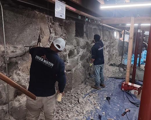 Workers clearing debris in a basement or crawl space with exposed rock walls and construction tools.