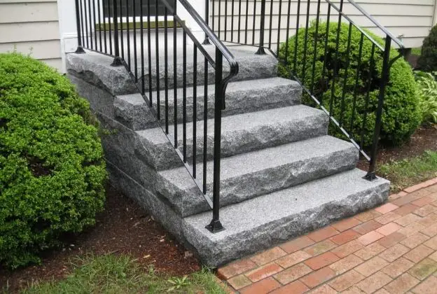 Gray stone front steps with black railings leading to a house, bordered by green shrubs and a brick walkway