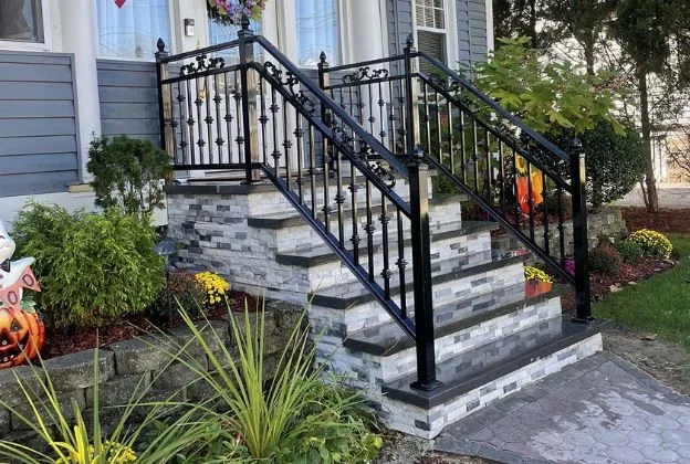 Front steps with black railings and stone trim leading to a gray house, with fall decorations and shrubs.
