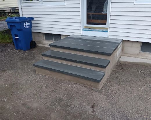 Back steps with dark treads beside a house, next to a blue recycling bin.