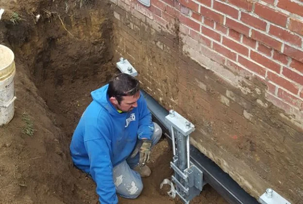 Worker in blue jacket kneeling beside a brick foundation, installing a metal support bracket.
