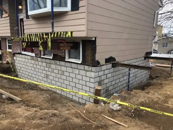 House corner with cinderblock foundation wall under construction, surrounded by caution tape and bare dirt