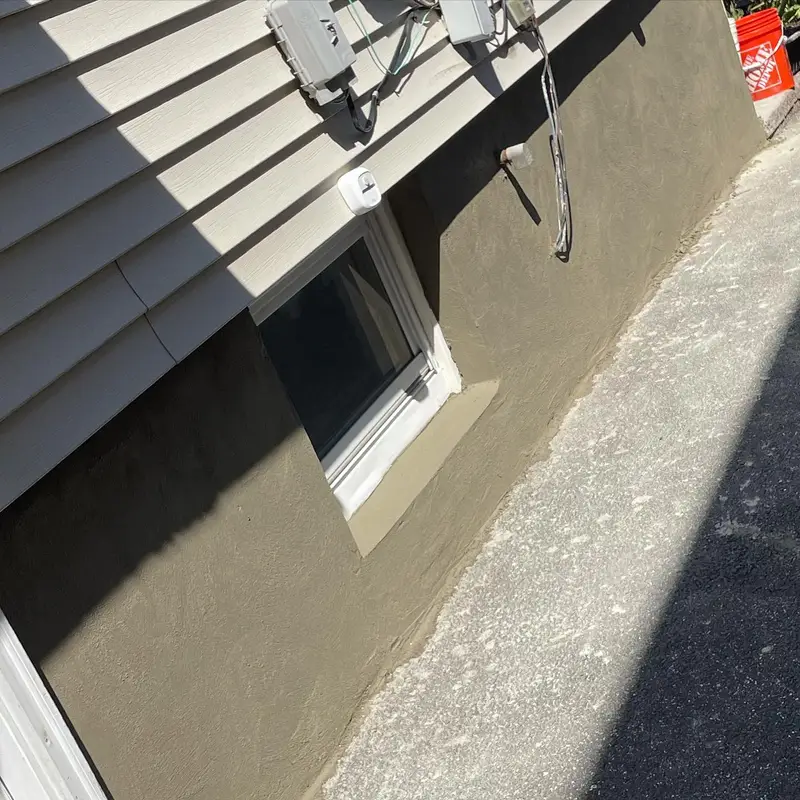 Basement window in beige house exterior beside utilities and a concrete walkway in sunlight