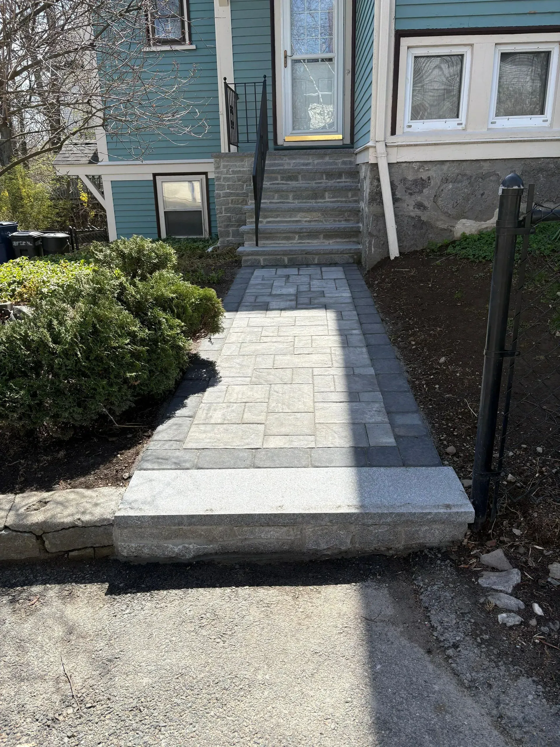 Stone walkway leading to a front door of a house, bordered by bushes and a black fence.
