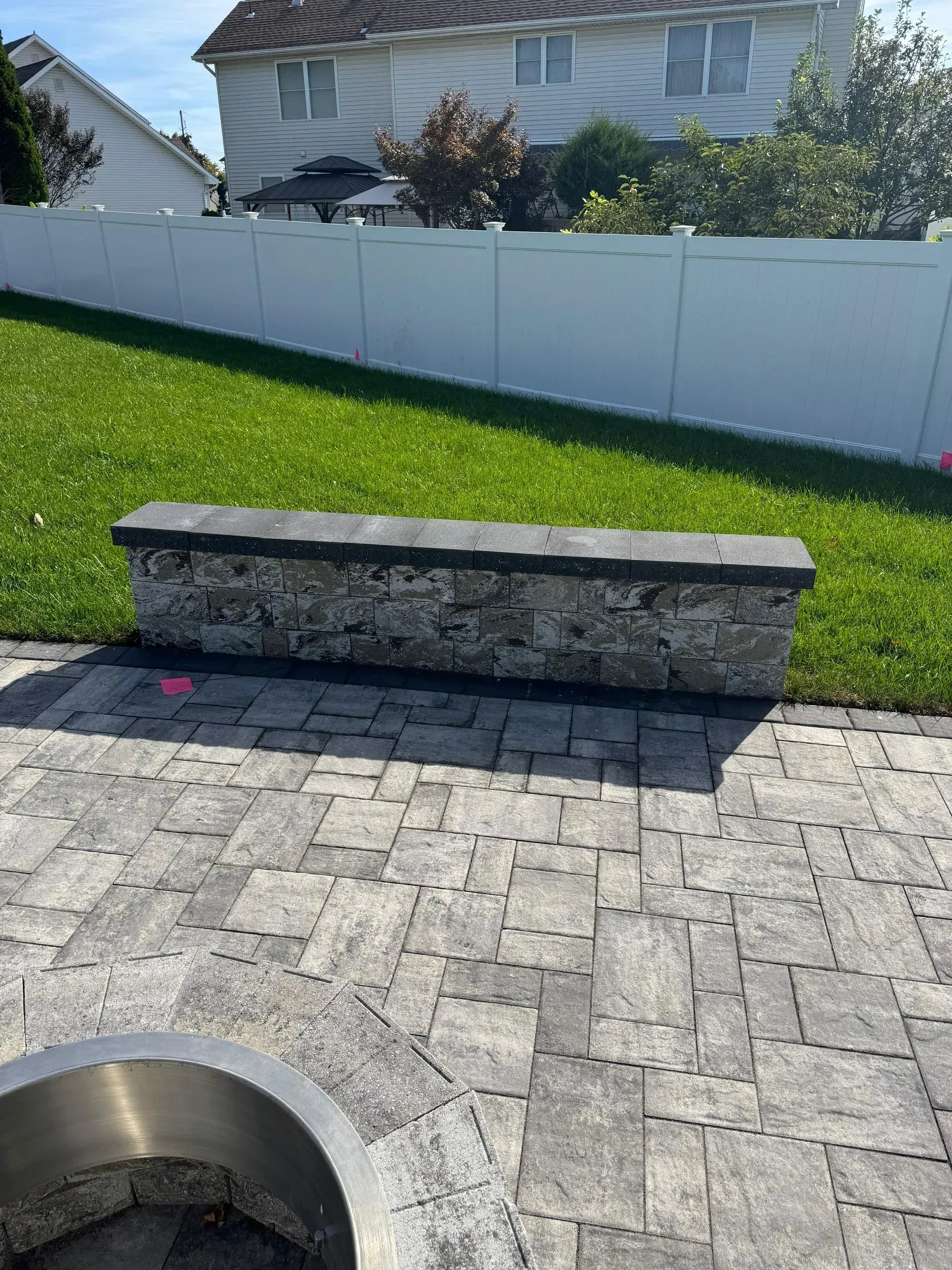 Stone bench on a brick patio beside a green lawn and white fence in a backyard.
