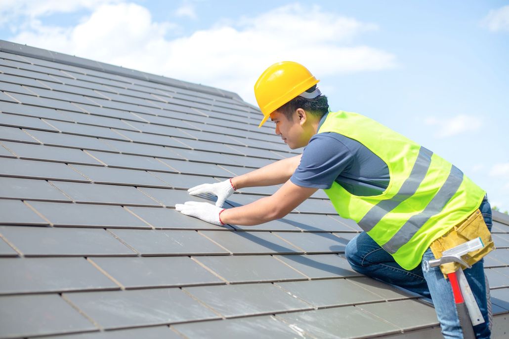 Worker in yellow hard hat and safety vest installing roof shingles on a sloped rooftop.