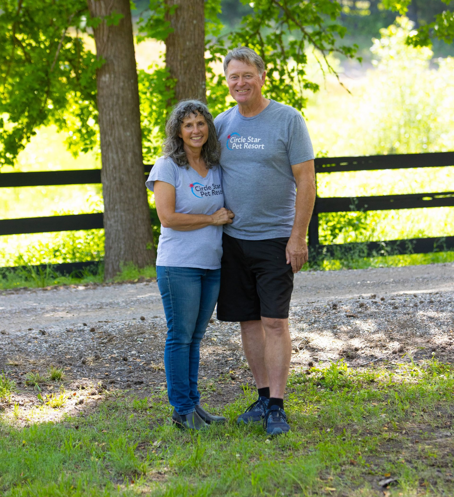 Couple standing outdoors near a fence and trees. Woman in jeans and t-shirt; man in shorts and a t-shirt.