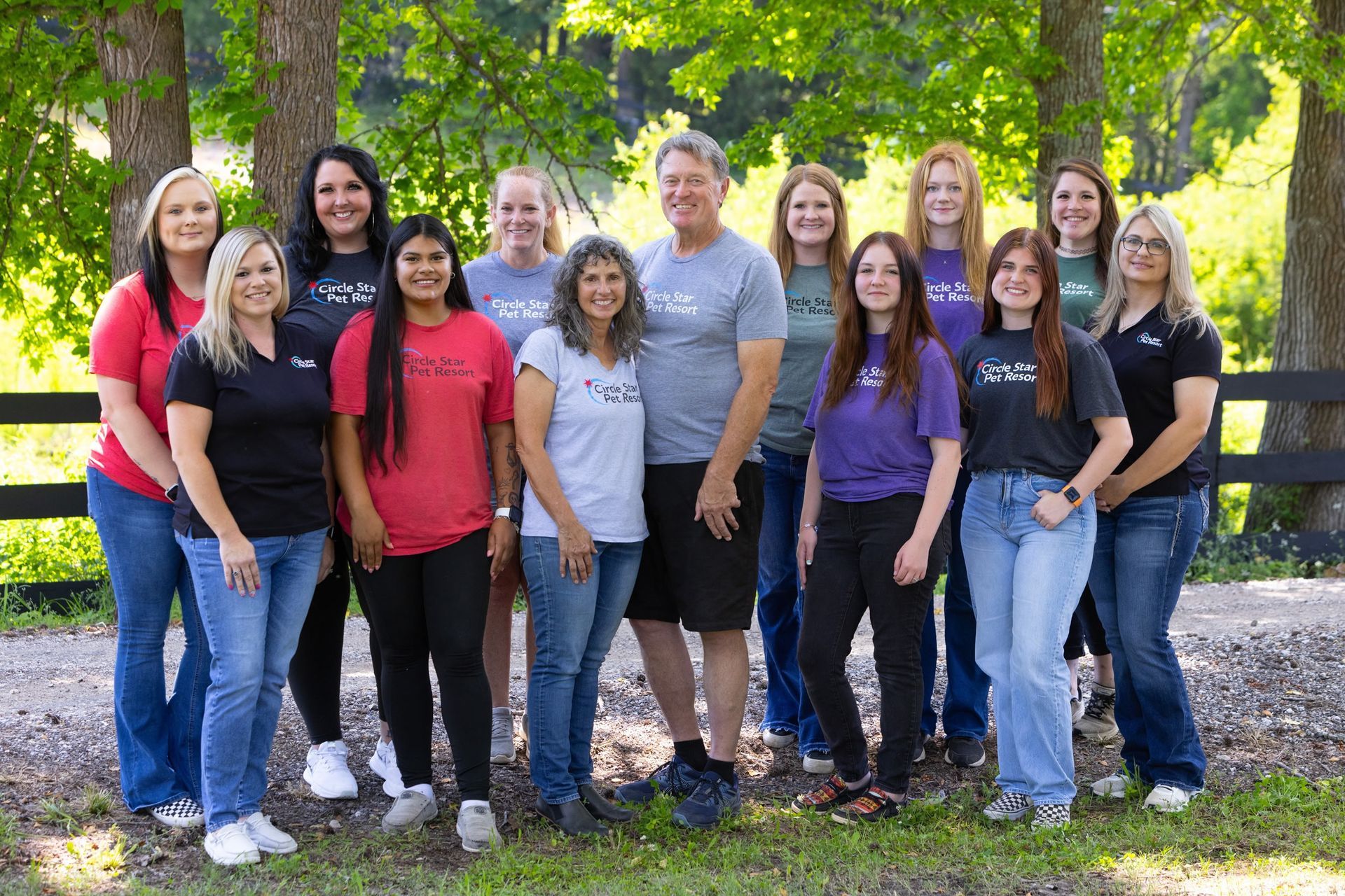 Group of people wearing t-shirts, posing outside, smiling. Green foliage, fence, trees visible.