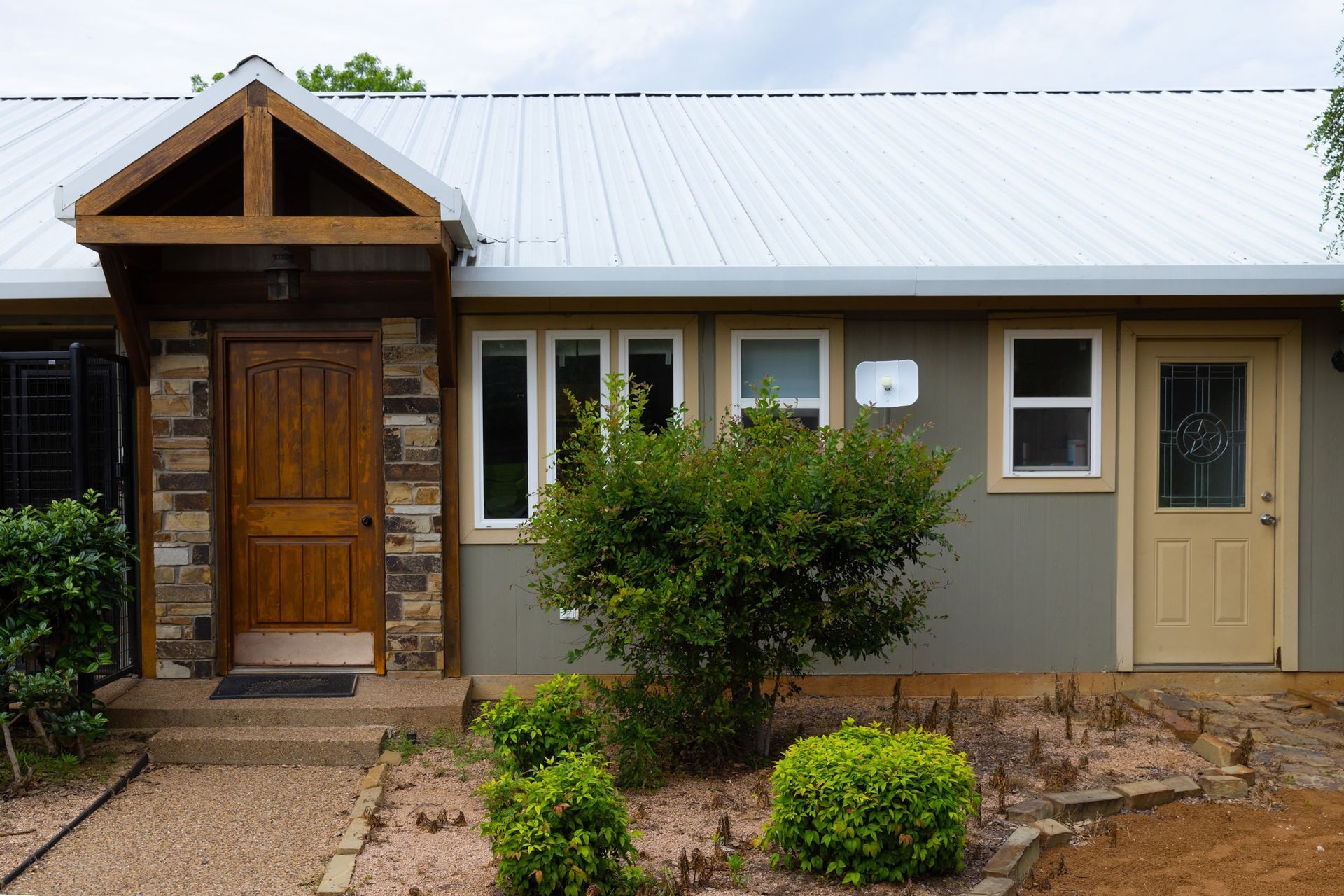 Small house with brown door, stone accents, and metal roof. Green siding, white trim, and small bushes.