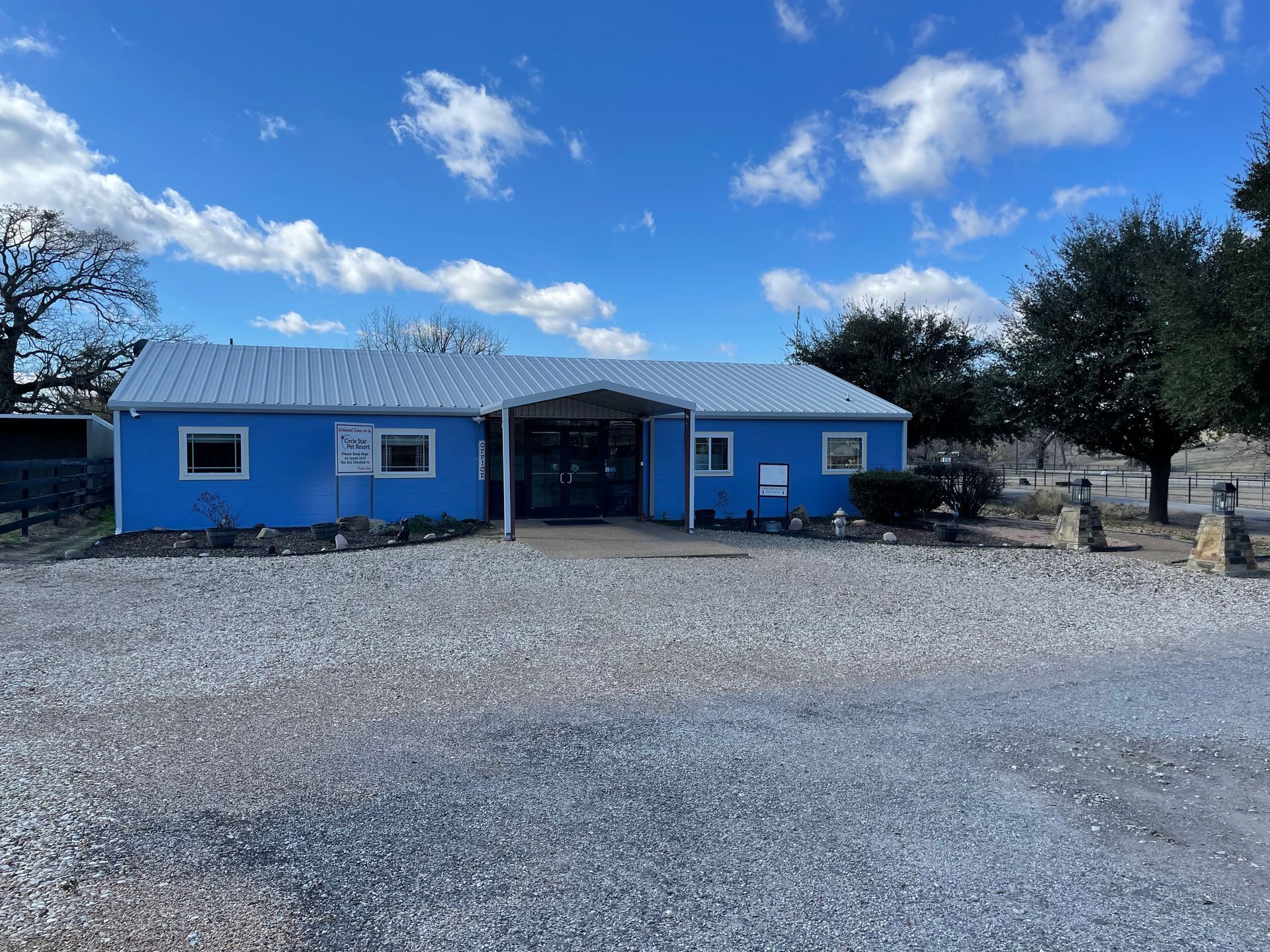 Blue building with a metal roof, gravel lot, and trees under a cloudy blue sky.
