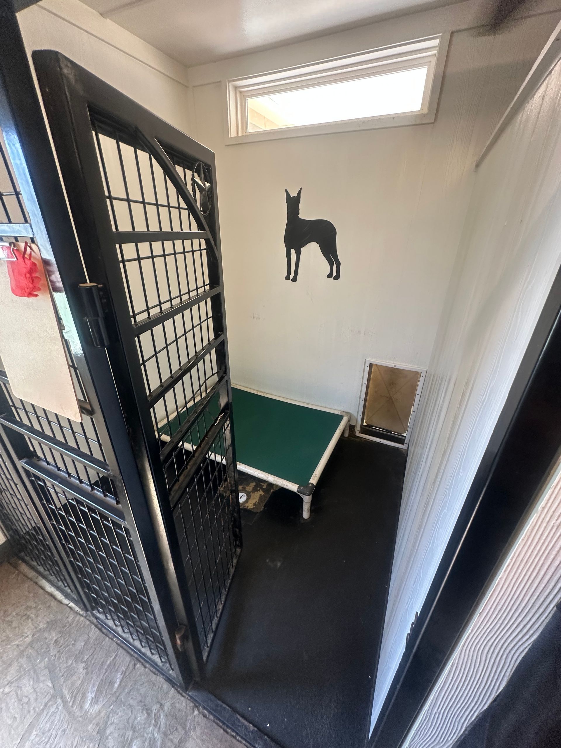 Dog kennel interior with black gate, bed, and dog silhouette on wall.