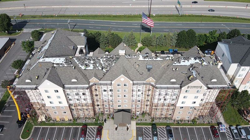 Aerial view of a multi-story building with a partially replaced roof. Cars are parked in the front. An American flag flies.