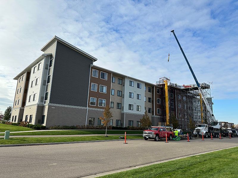 Multi-story building under construction with a crane and trucks, on a green lawn with a blue sky.