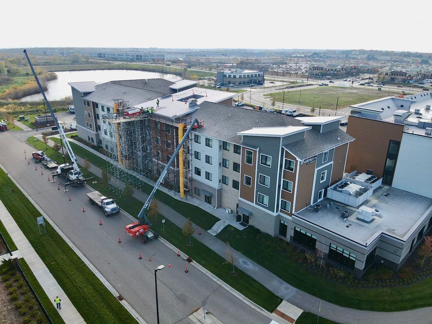 Aerial view of a multistory building under construction, with cranes, trucks, and workers on the roof, near a lake.