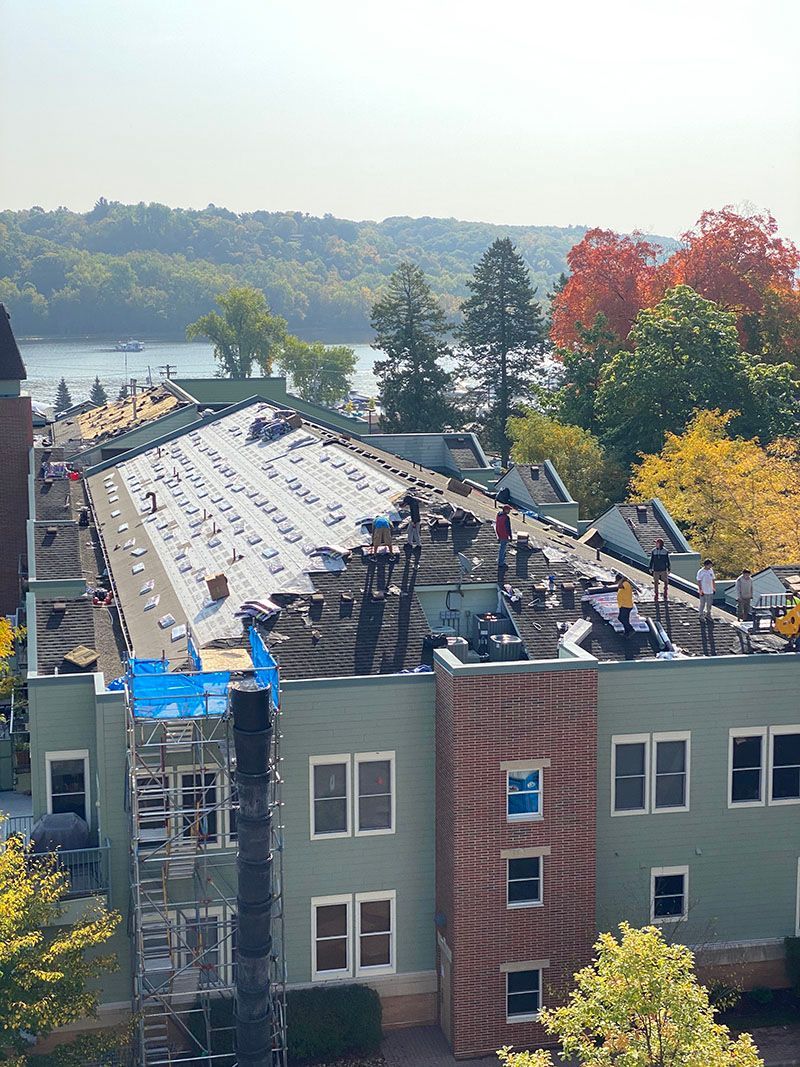 Workers on a rooftop during a sunny day, with a lake and trees in the background. Building has green and brick sections.