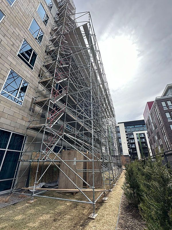 Scaffolding against a building facade. The building has windows and textured gray blocks. Cloudy sky above.
