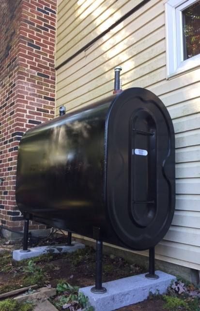 Black oil tank outside a house, against beige siding and brick chimney.