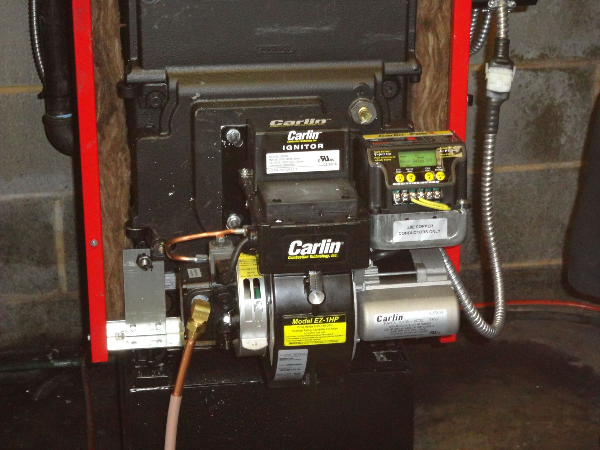 Close-up of a Carlin brand oil burner attached to a furnace, against a cinder block wall.