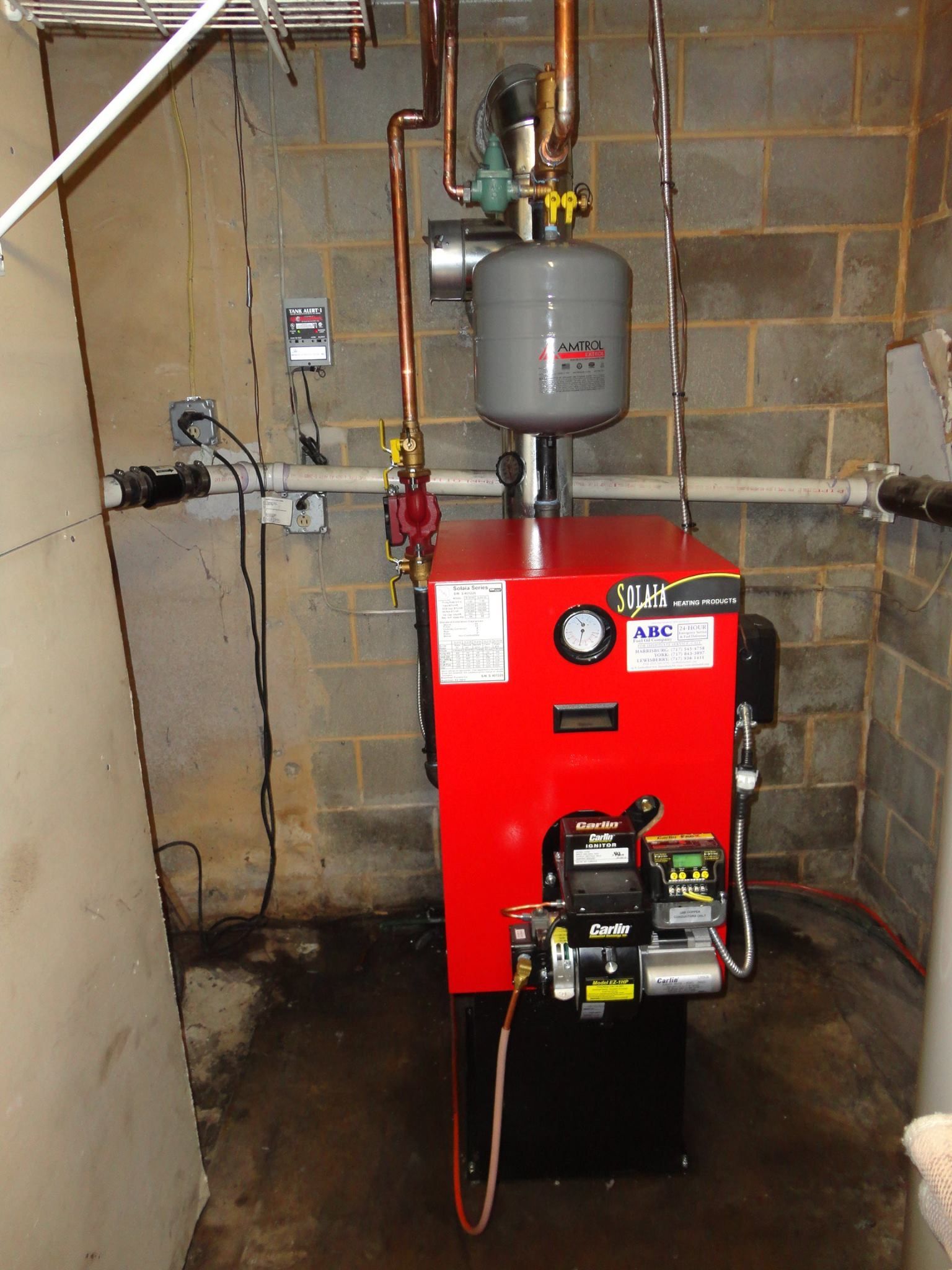 Red heating oil boiler with copper pipes, gray expansion tank, and brick wall background.