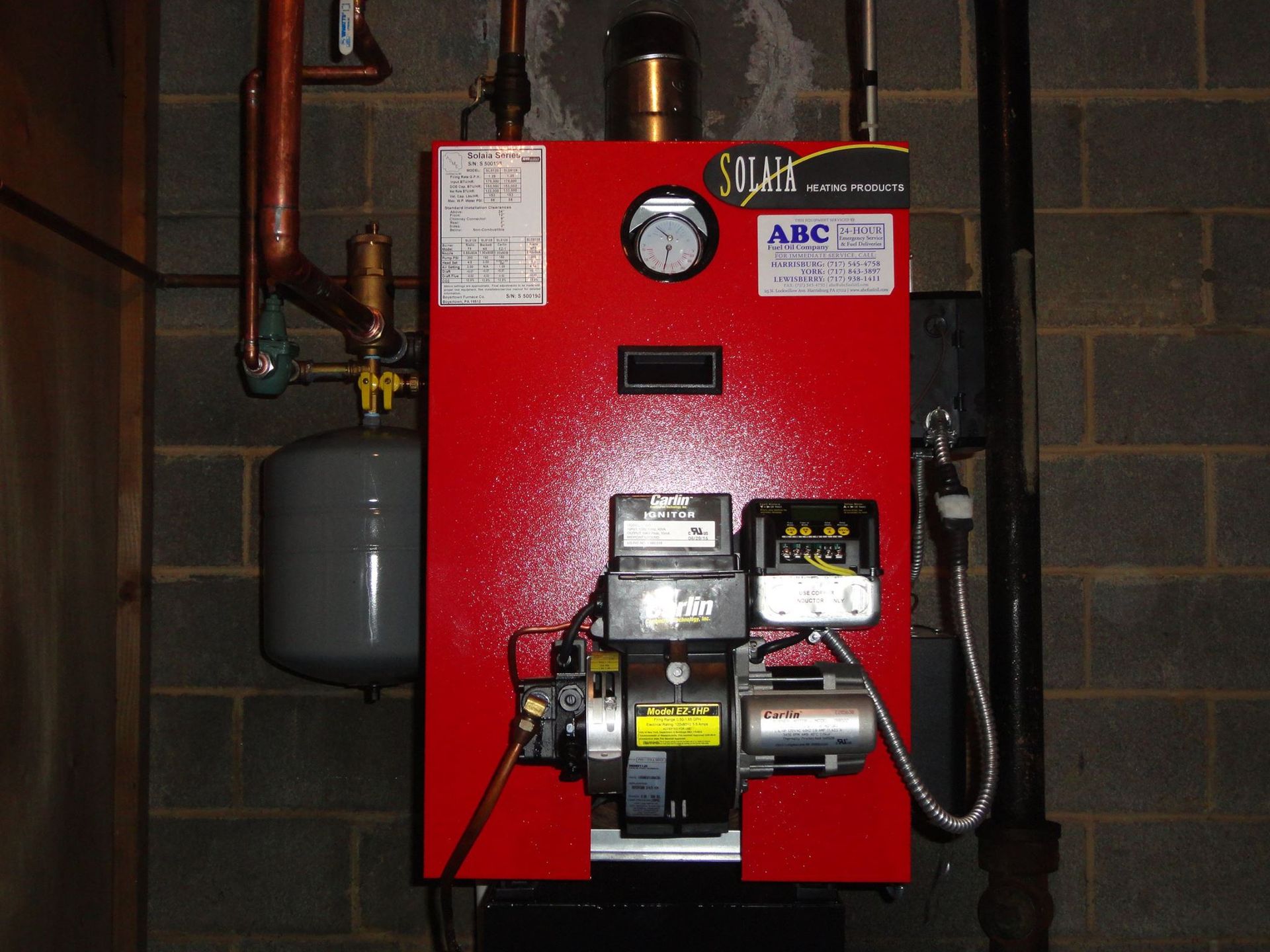 Red oil furnace mounted on a cinder block wall, with copper piping and a gray expansion tank.