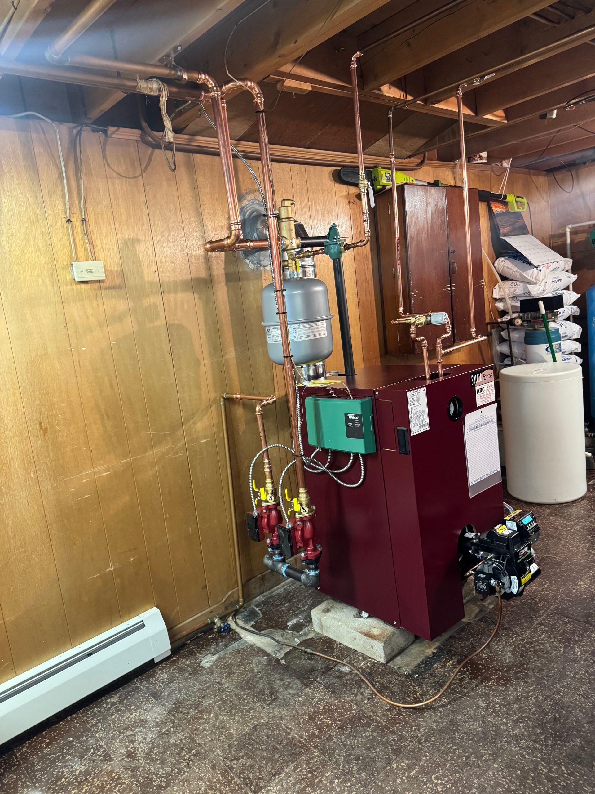 A boiler system with copper pipes, a green pump, and a cylindrical pressure tank in a basement setting.