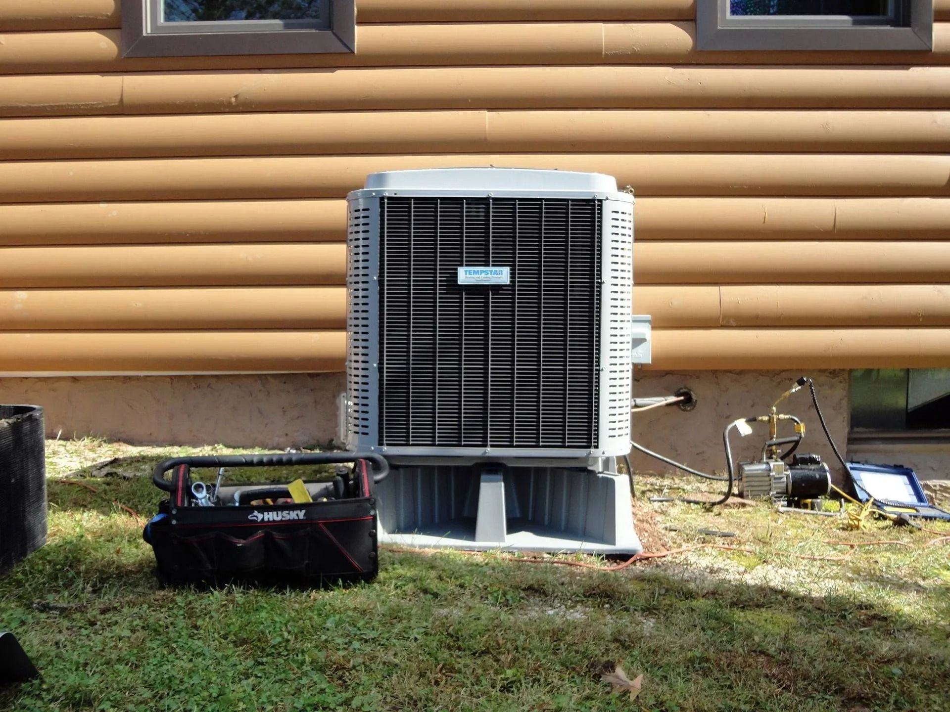 Air conditioning unit against a wood-sided house with a tool bag and equipment on the ground.