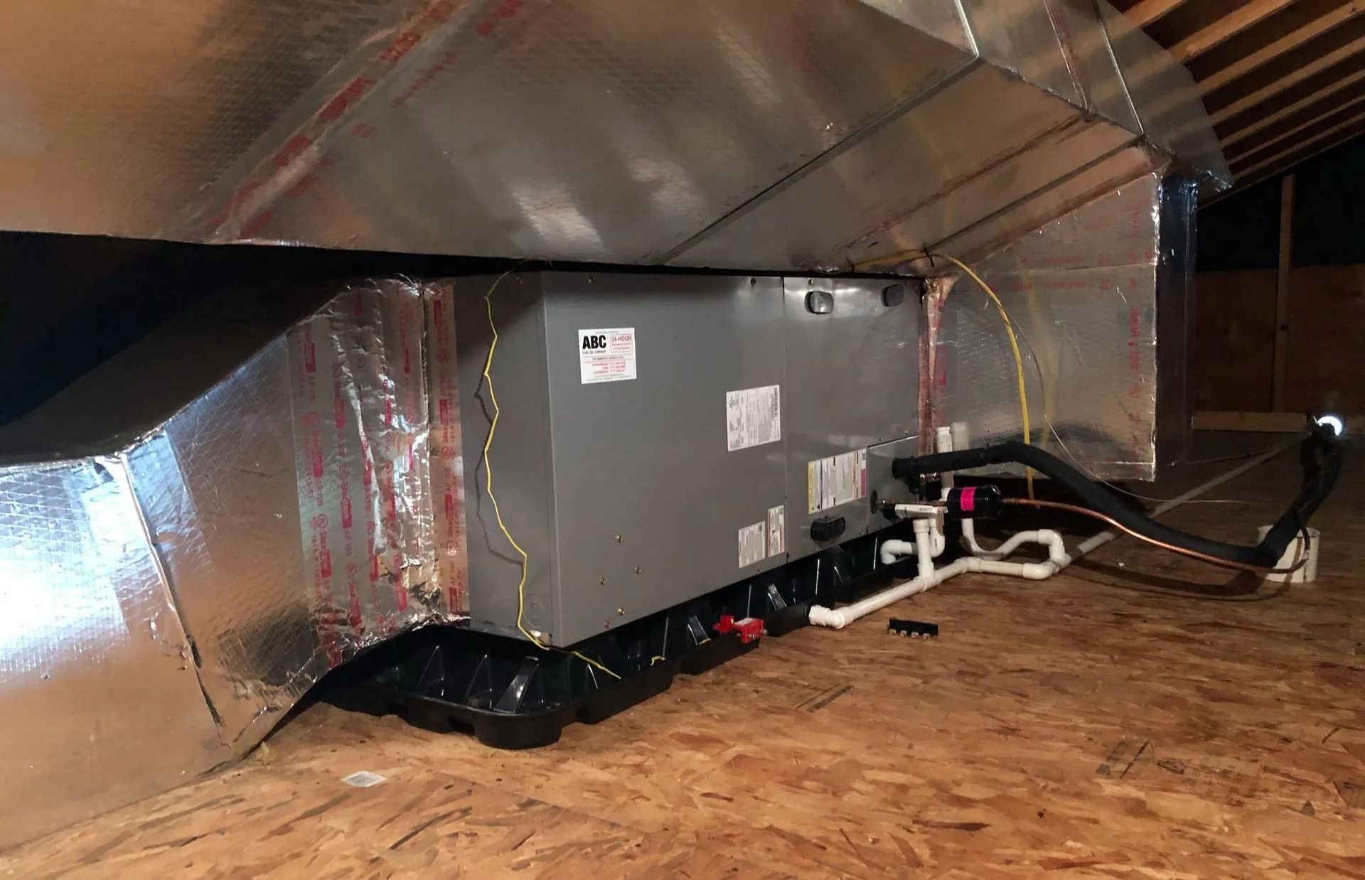HVAC unit installed in an attic. Silver insulated ductwork, grey unit, black base, wood flooring.