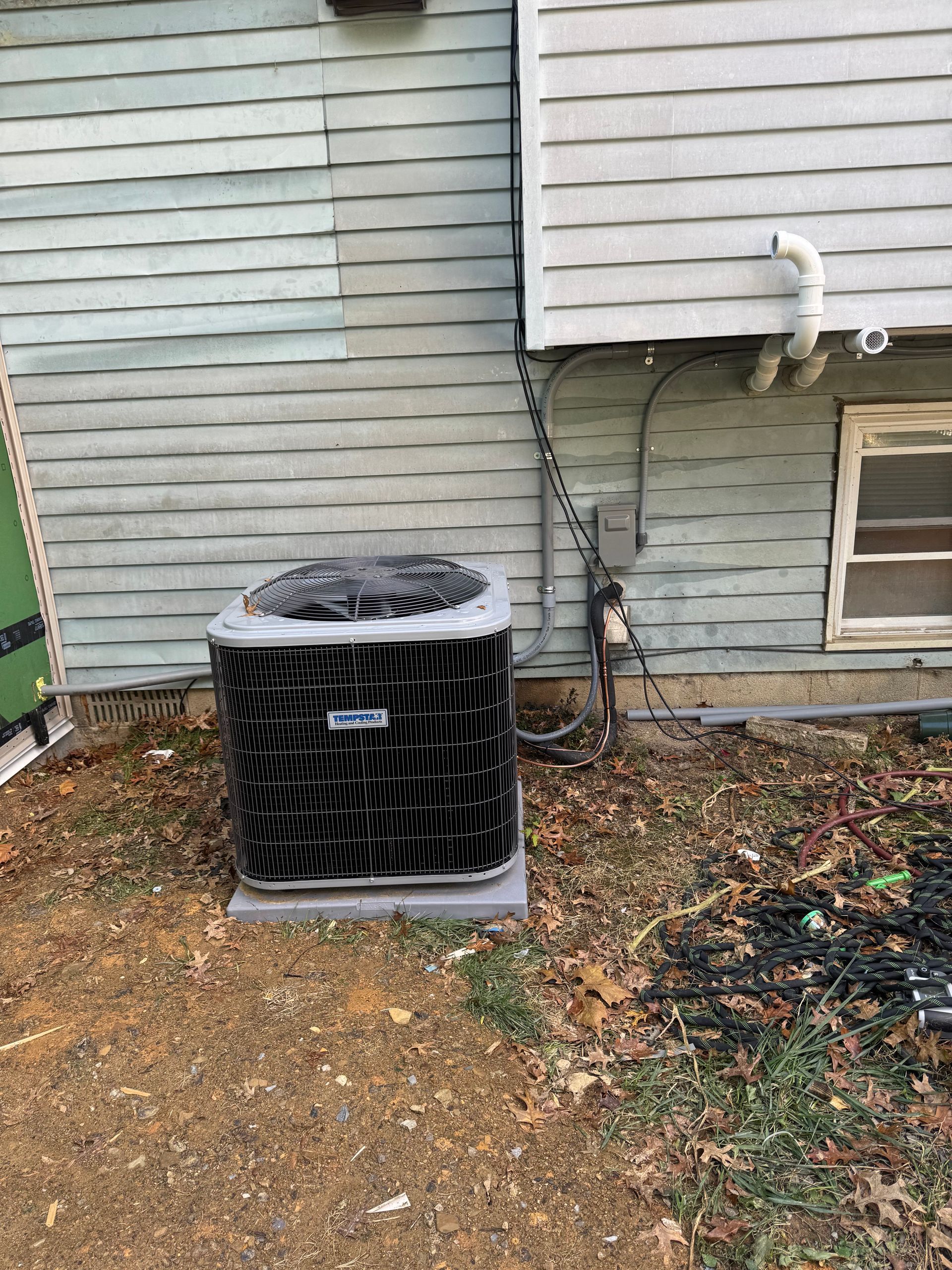 Air conditioning unit against a light blue siding building, surrounded by leaves and dirt.