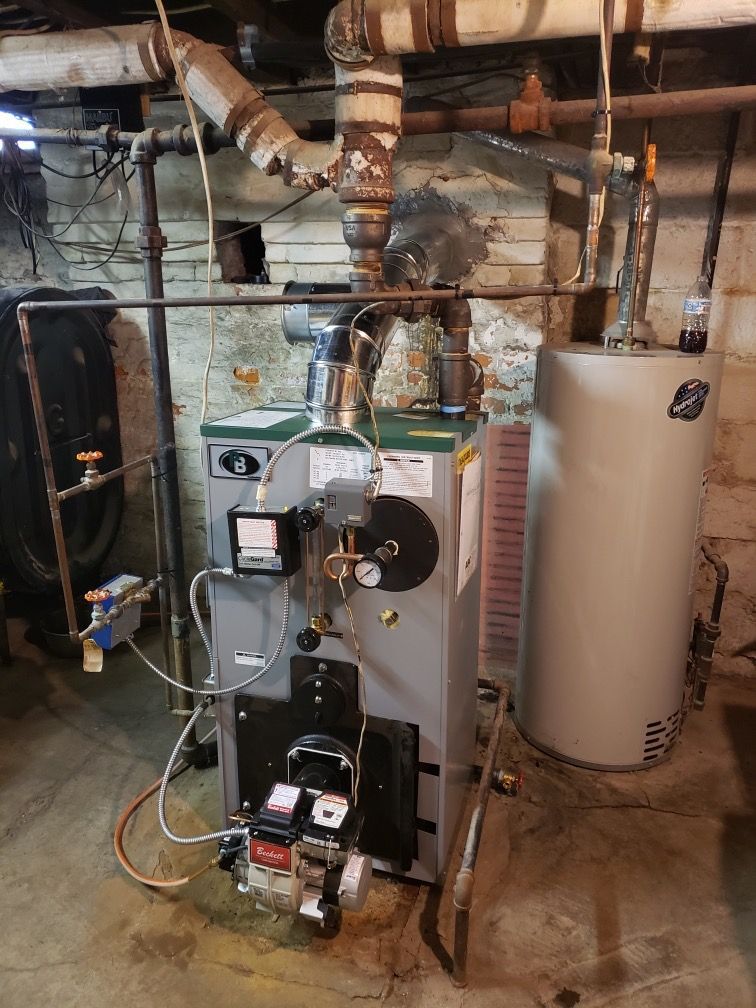 Furnace and water heater in a basement. Grey furnace with piping, connected to a vertical water heater.