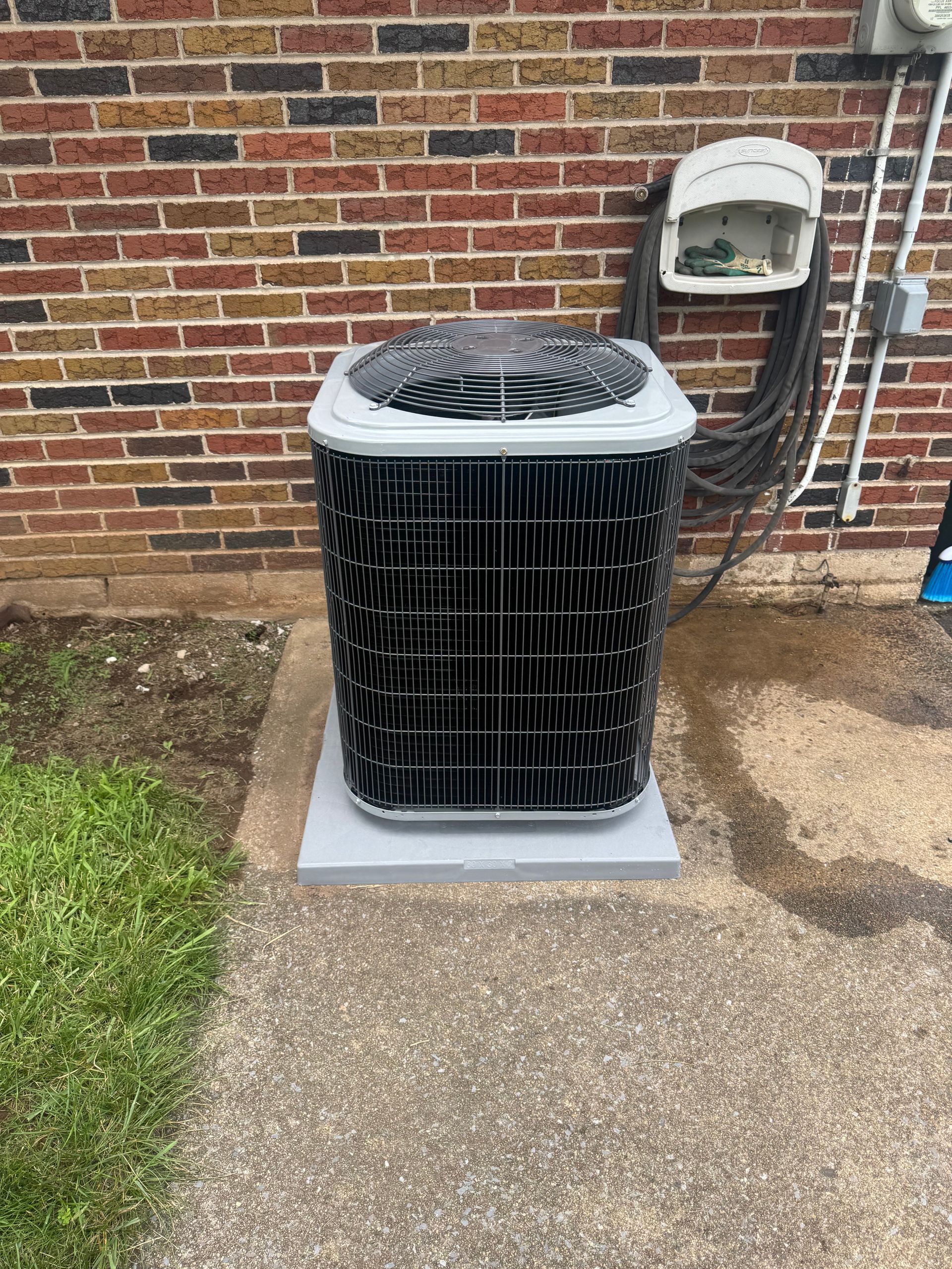 AC unit on a concrete pad next to a brick wall. A garden hose hangs nearby.