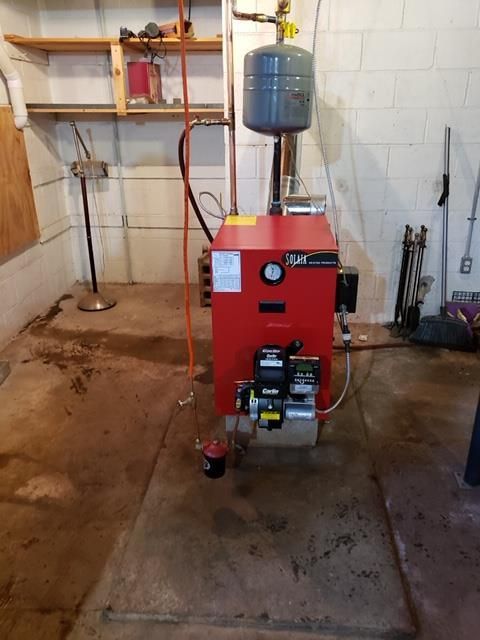 Red oil burner in a basement, with an expansion tank, pipes, and concrete floor.