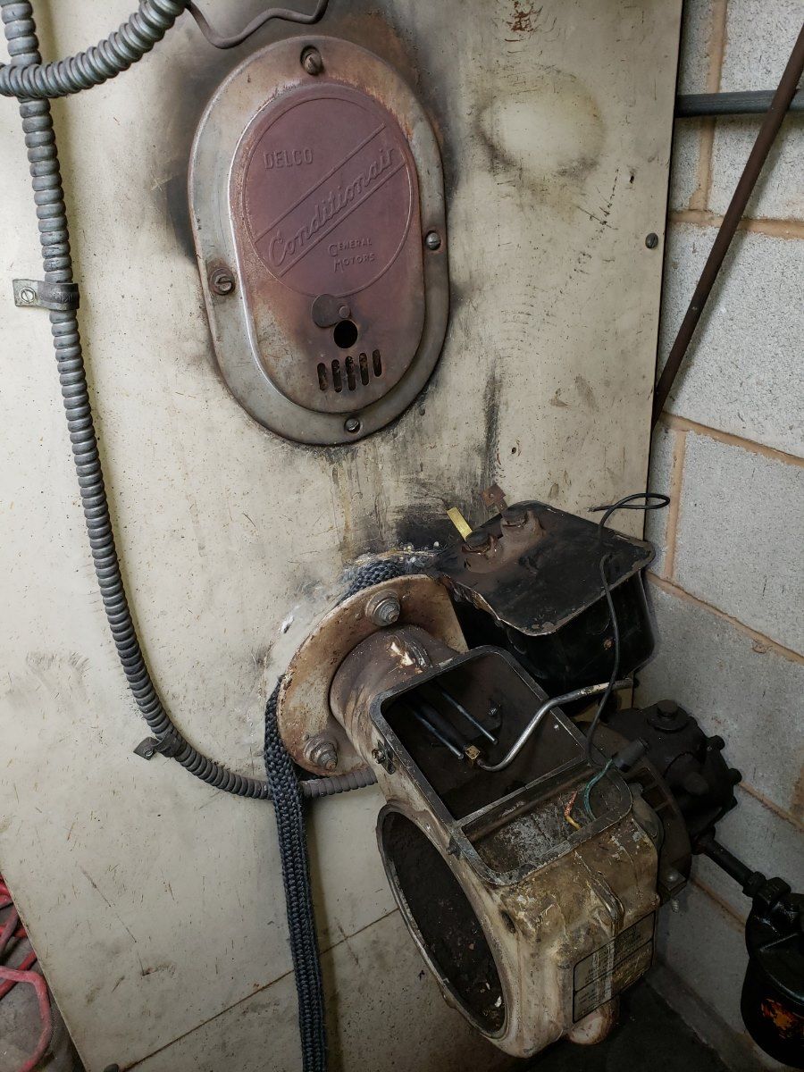 Old oil furnace with oval access panel and attached burner; black soot stains the wall.