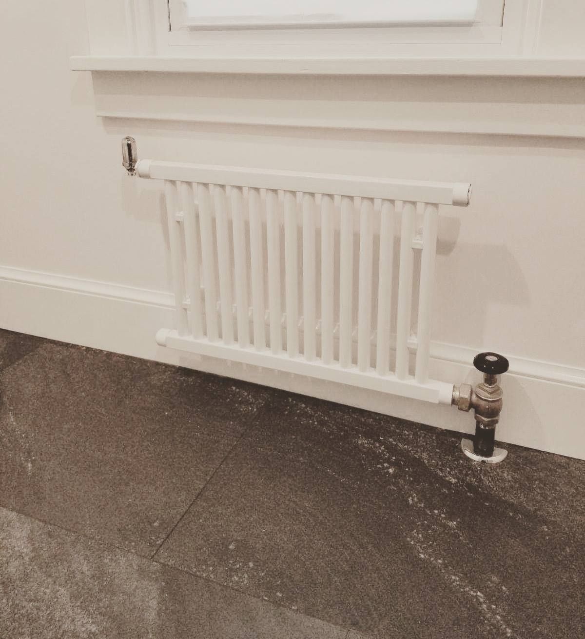 White radiator mounted on a wall below a window. A valve is connected to a pipe emerging from the floor.
