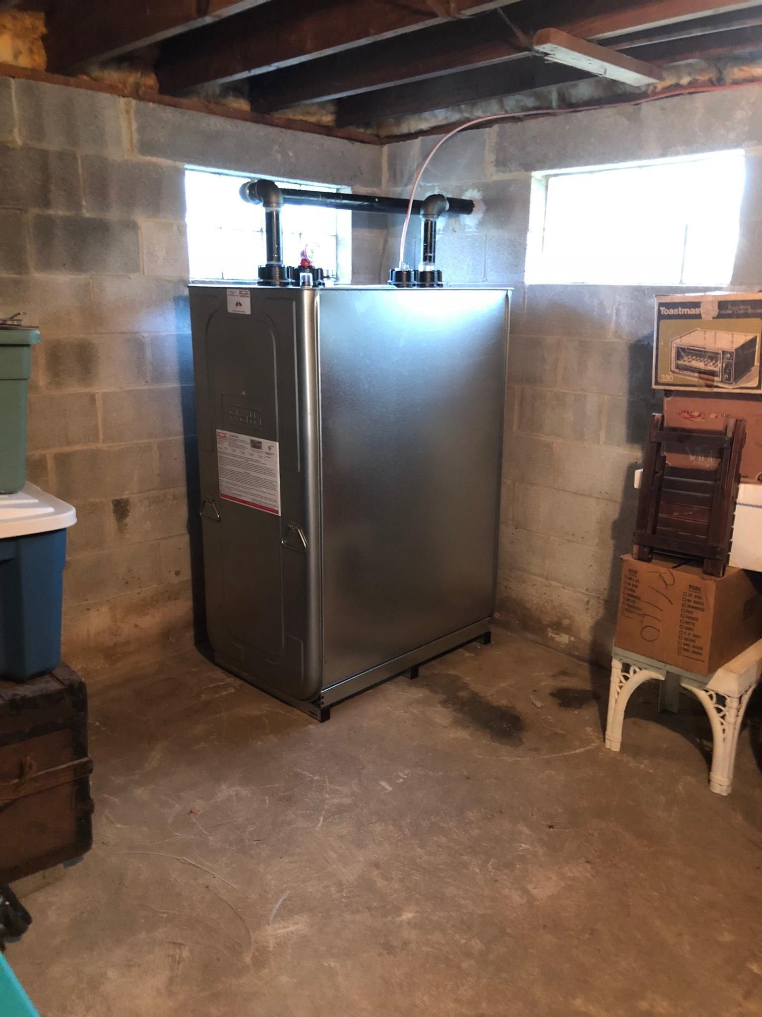 A large, silver heating unit in a basement. Beside it, are stacked boxes and a chair.