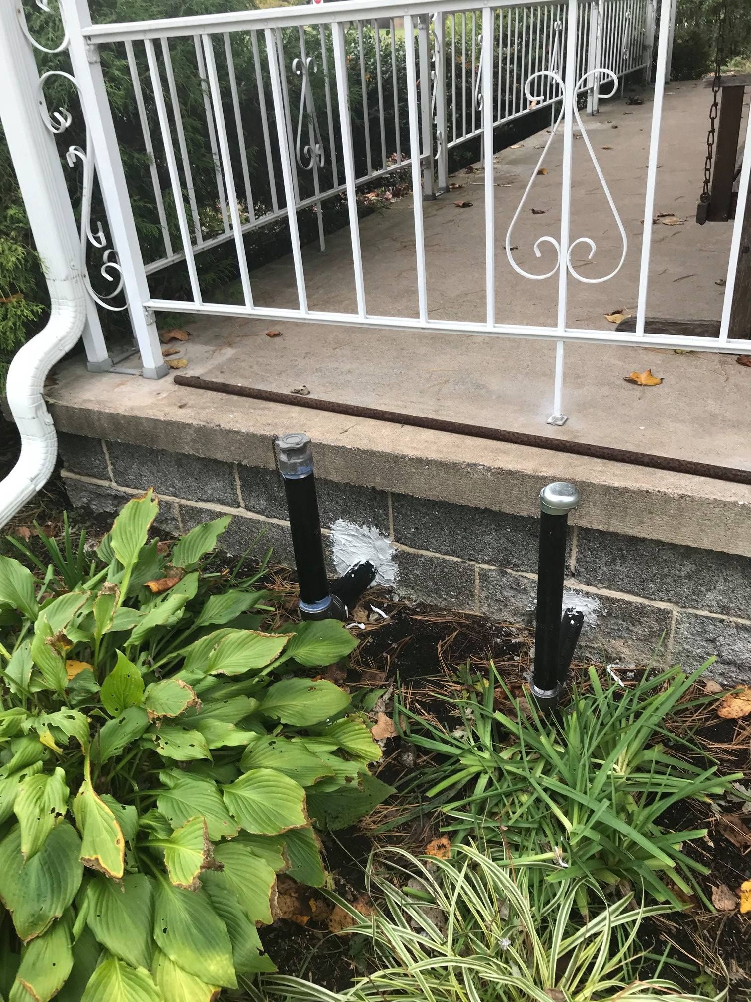 Two black pipes emerge from a concrete foundation, next to green plants and a white railing on a porch.