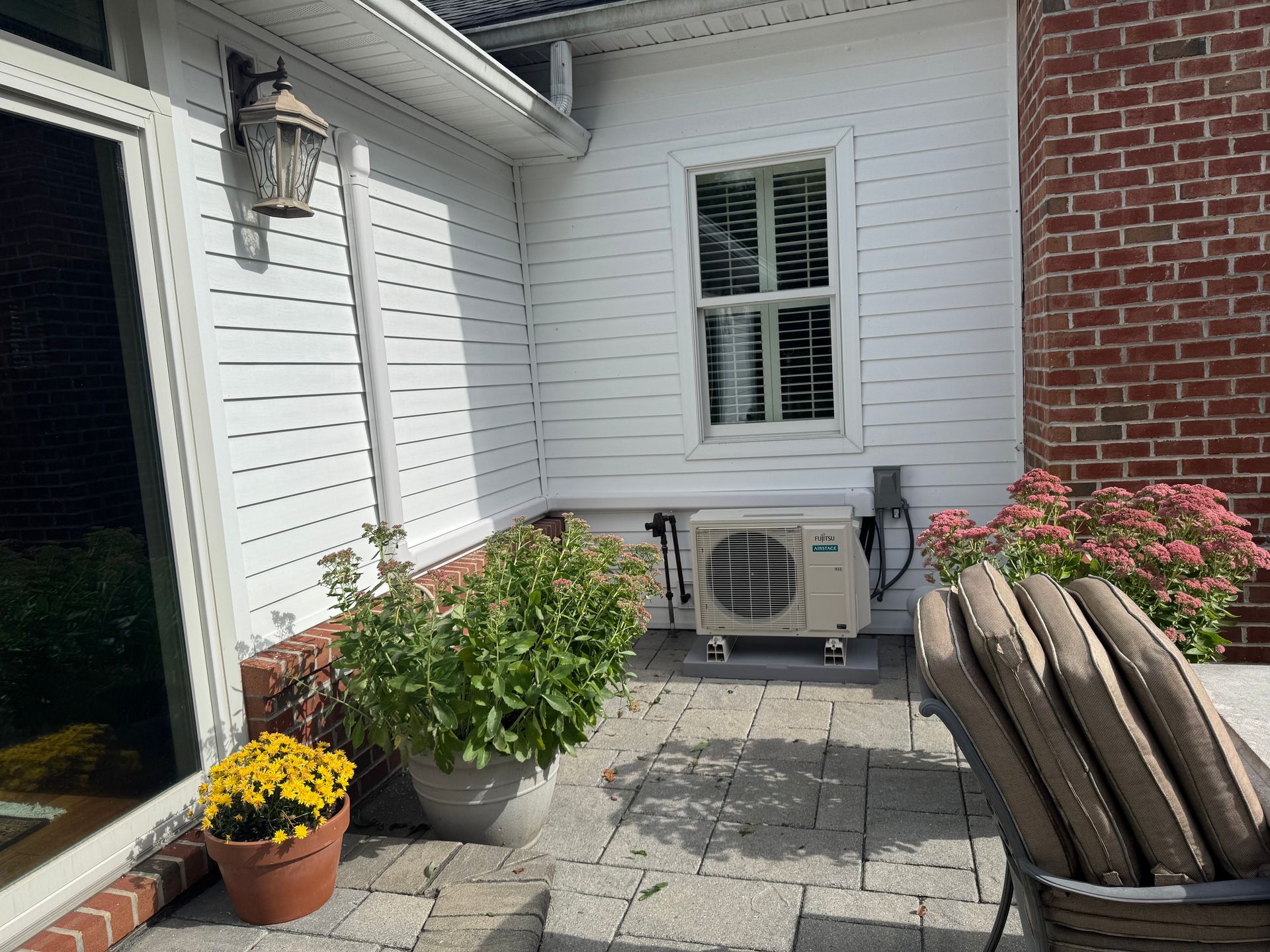 White house exterior with air conditioning unit, window, and potted plants on a brick patio.